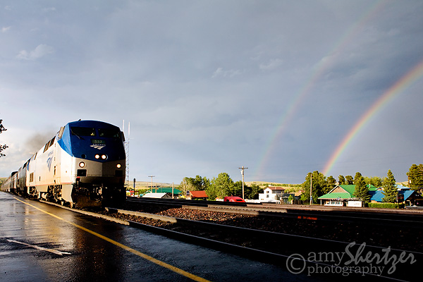 An Amtrak train arrives at East Glacier, MT with a double rainbow lighting up the sky