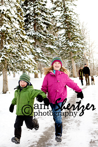 Young kids run through the snow with their parents watching in the background.